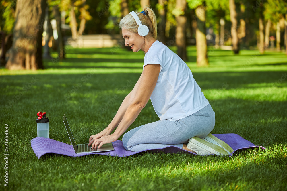 Fototapeta premium Joyful woman in headphones using notebook in park
