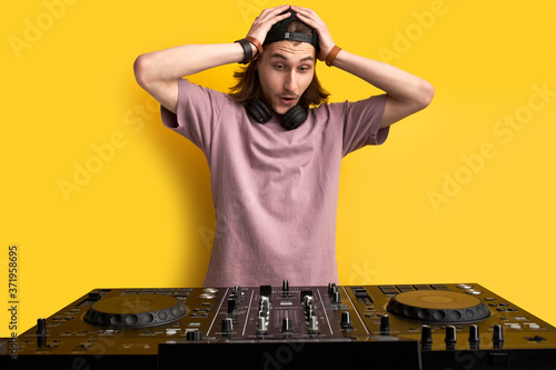 dj man shocked after seeing vinyl turntable equipment, going to perform music on it, hold hands on head, surprised. isolated yellow background