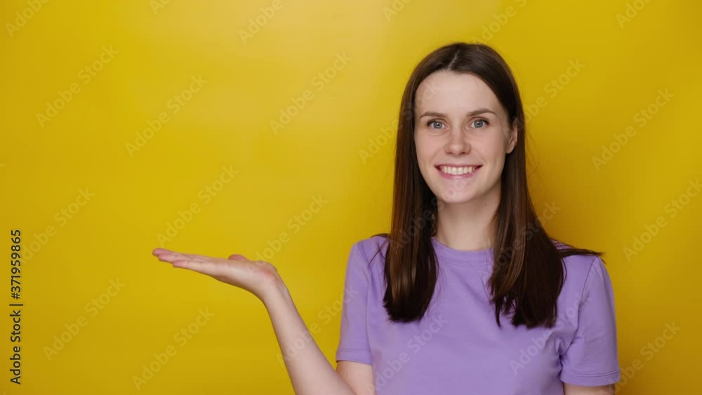 Smiling cute woman looks pleasant to her palm hand, showing something in her hand, wears purple t-shirt, isolated on yellow studio background with copy space. People sincere emotions concept