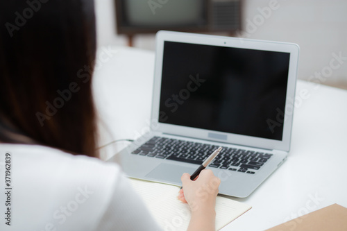 Young asian woman writing on notebook for planning working and using laptop computer blank screen on desk at home, girl notes, female study and learning, business and communication concept.