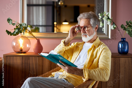 intelligent mature man read book at home, enjoy free time alone, sit on chair, wearing eyeglasses for better reading