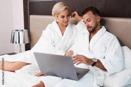 beautiful caucasian couple lie on bed with laptop, discuss something, wearing white bathrobe. lovely woman and man in hotel