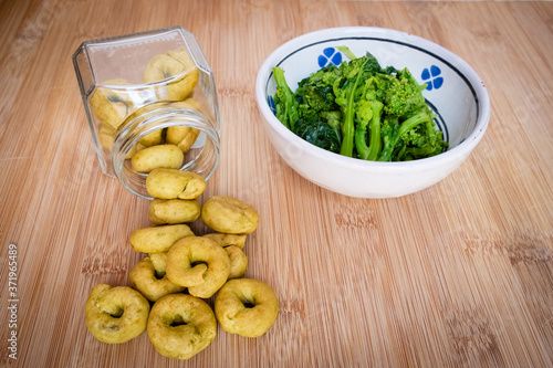 Homemade rapini flavour taralli crackers poured from a glass jar next to a bowl full of boiled rapini. Traditional snack food of Puglia