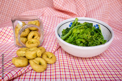 Homemade rapini flavour taralli crackers poured from a glass jar next to a bowl full of boiled rapini. Traditional snack food of Puglia