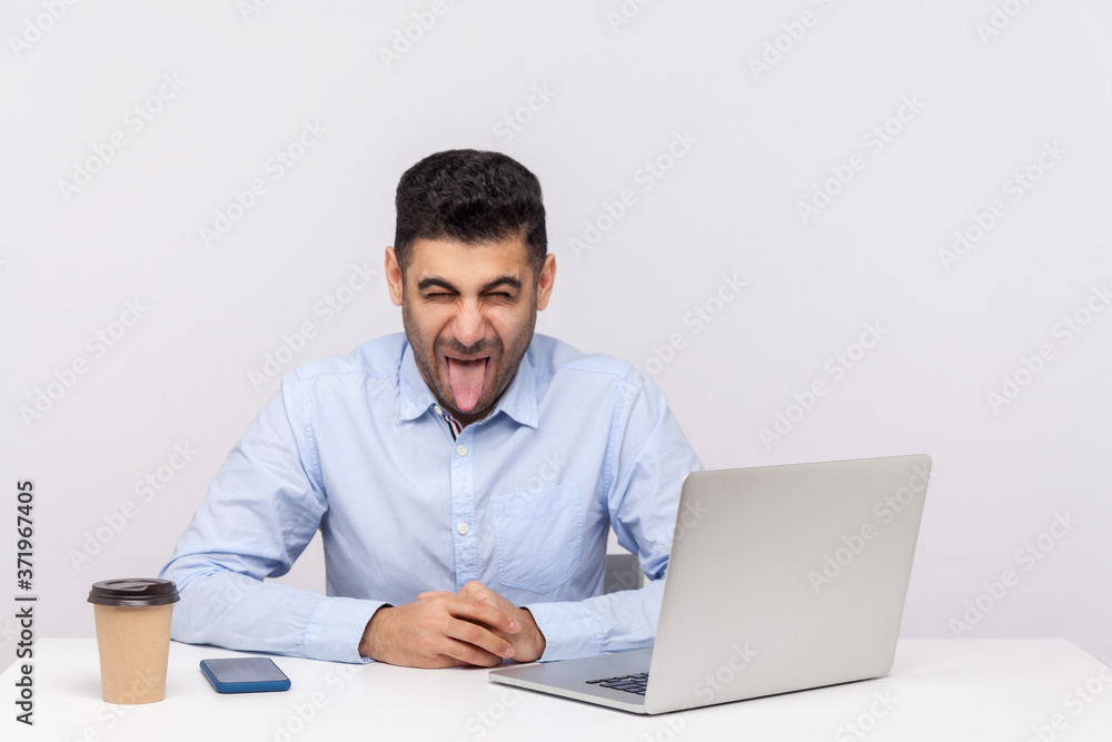 Crazy funny businessman sitting office workplace with laptop on desk, showing his tongue, looking at camera with naughty disobedient expression. indoor studio shot isolated on white background Crazy funny businessman sitting office workplace with laptop on desk, showing his tongue, looking at camera with naughty disobedient expression. indoor studio shot isolated on white background