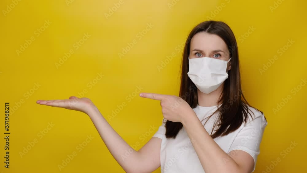 Young woman wears respirator white mask to protect herself from coronavirus, demonstrates empty copy space for promo text, isolated over yellow studio background. Pandemic spreading virus concept Young woman wears respirator white mask to protect herself from coronavirus, demonstrates empty copy space for promo text, isolated over yellow studio background. Pandemic spreading virus concept