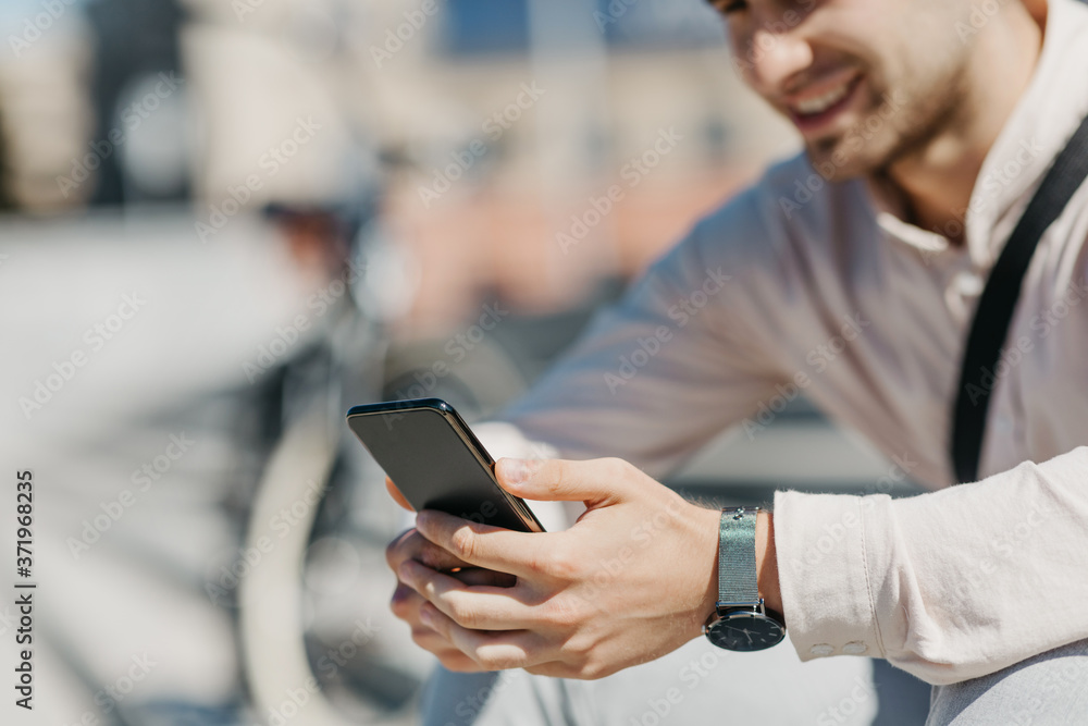 Online in city. Smiling attractive guy in shirt sits on street and looks at smartphone Online in city. Smiling attractive guy in shirt sits on street and looks at smartphone