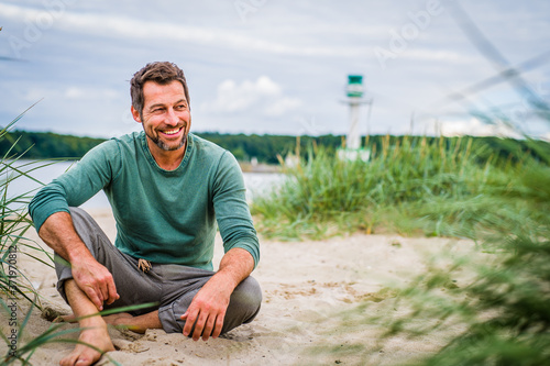 Mann im Besten Alter und sportiv leger gekleidet sitzt an Strand und Harmoniert mit dem Hintergrund