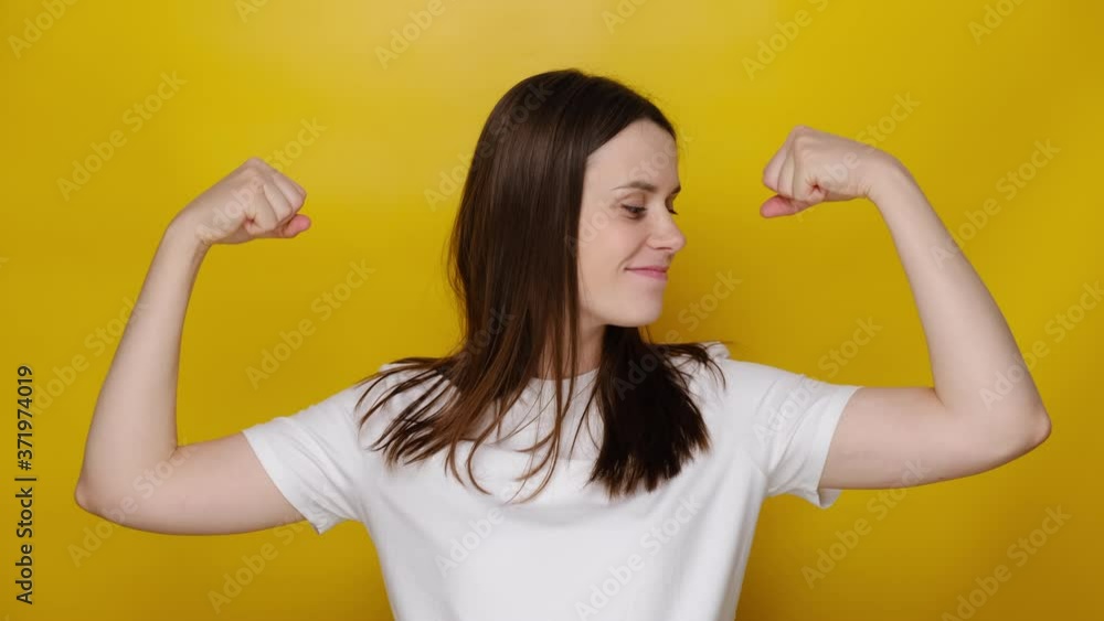 Portrait of attractive young woman showing biceps and looking confident, feeling power strength to fight for female rights, isolated on yellow studio background. I am strong and independent!