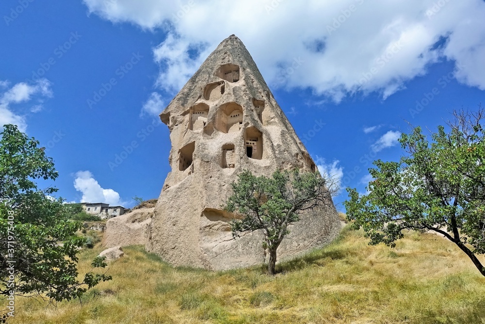 A strange ancient rock of Cappadocia against the backdrop of a bright blue sky. In the conical cliff, caves were cut down - the dwellings of ancient people. Around the yellowed grass, trees. Turkey.