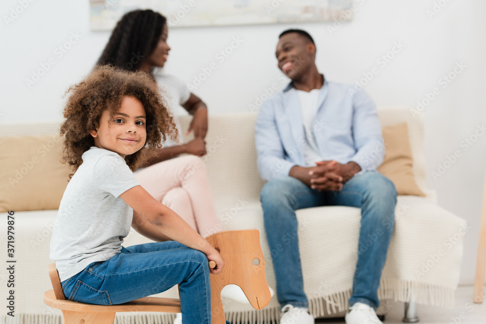 selective focus of curly african american kid sitting on rocking horse and looking at camera near parents selective focus of curly african american kid sitting on rocking horse and looking at camera near parents