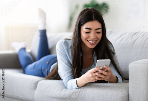 Useful App. Korean Girl Using Smartphone While Relaxing On Sofa At Home