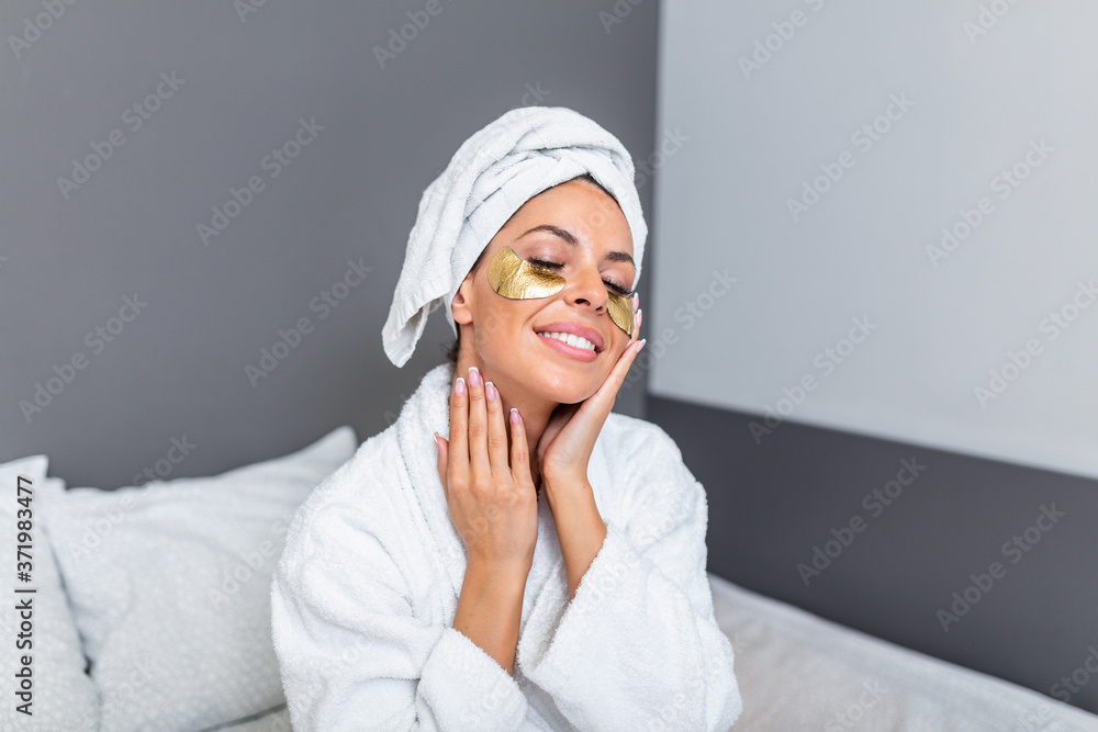 Young woman face with Gold Cosmetics mask, Eye Patches and perfect skin wearing bath towel and bathrobe, sitting on the bed