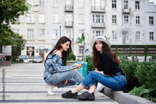 Two young brunette girls, wearing casual jeans clothes, with mint luggage, sitting on pavement border, checking phone in front of historical building in town. City sightseeing tour.Tourism lifestyle.