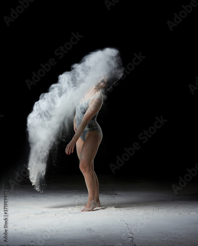 beautiful caucasian woman in a black bodysuit with a sports figure is dancing in a white cloud of flour
