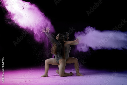 two beautiful young caucasian women in black bodysuits with a sports figure are dancing in a purple-blue cloud of flour on a black background