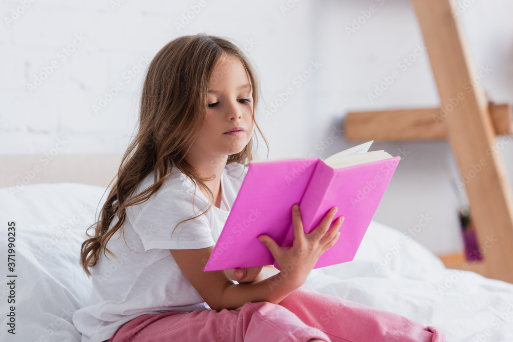 concentrated girl in pajamas sitting on bed and reading book concentrated girl in pajamas sitting on bed and reading book