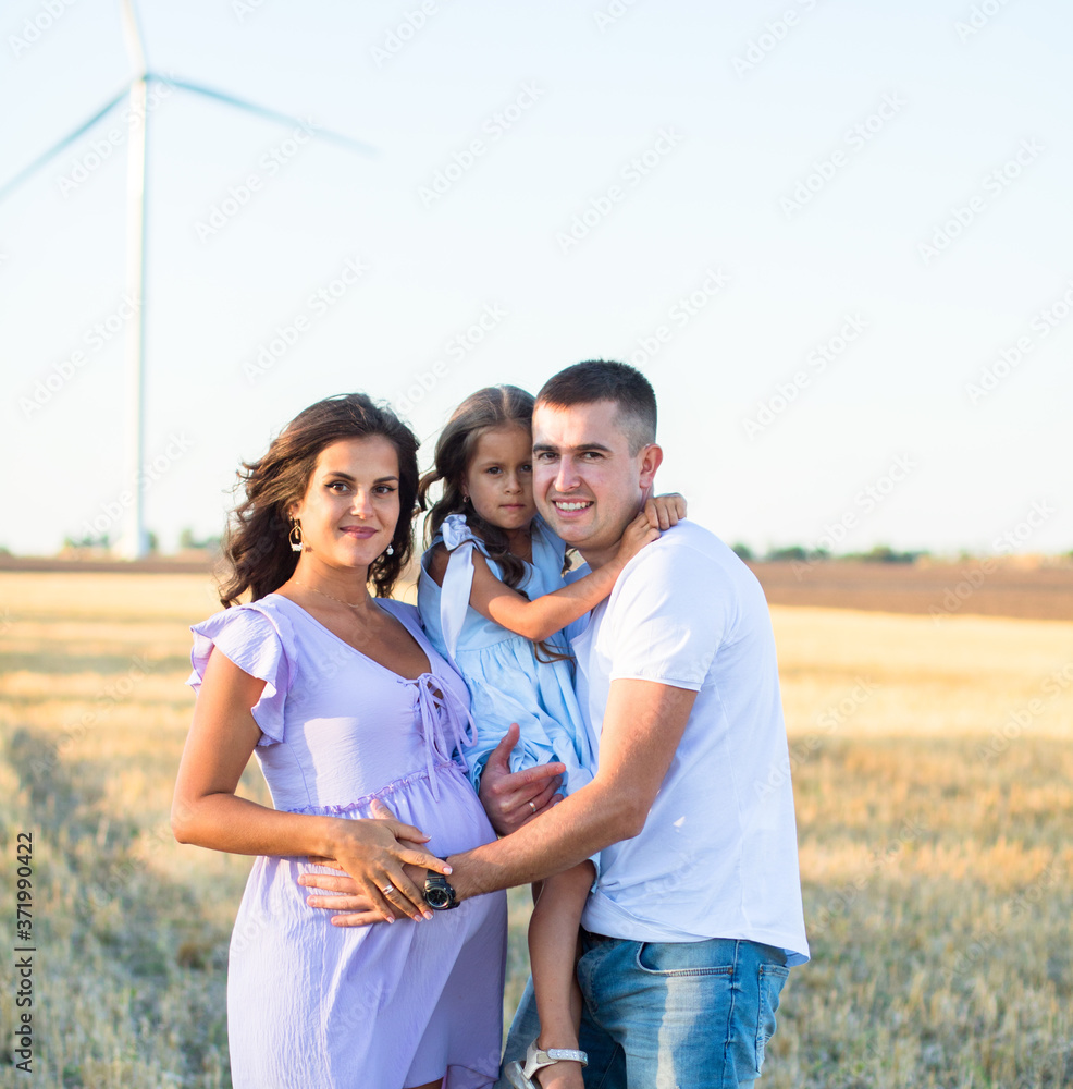 Obraz premium happy young family on the field, pregnant wife and a little girl hugs with father, wind turbines on the background