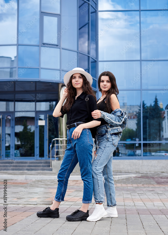 Two young brunette girls, wearing casual jeans attire, standing in front of blue modern glass building. Girlfriends traveling in city, holding backpacks, sightseeing. Active lifestyle concept.