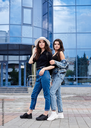 Two young brunette girls  wearing casual jeans attire  standing in front of blue modern glass building. Girlfriends traveling in city  holding backpacks  sightseeing. Active lifestyle concept.