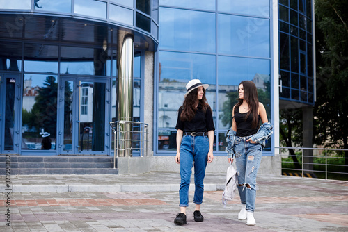 Two young brunette girls, wearing casual jeans attire, walking in front of blue modern glass building. Girlfriends traveling in city, holding backpacks, sightseeing. Active lifestyle concept.