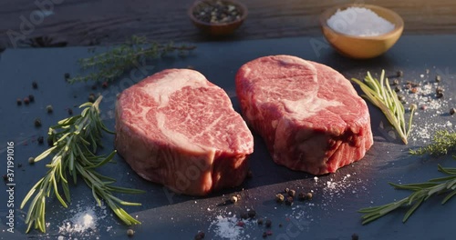 Two raw rib eye steaks with spices and herbs on a stone surface. The camera rotates slowly around the steaks.