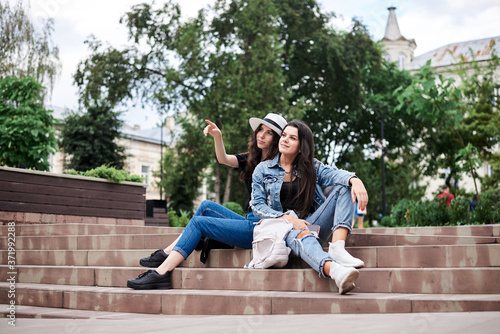 Two young brunette girls, wearing casual jeans clothes, holding small backpacks, sitting on stairway, resting, in front of historical building in town. City sightseeing tour. Traveling together.