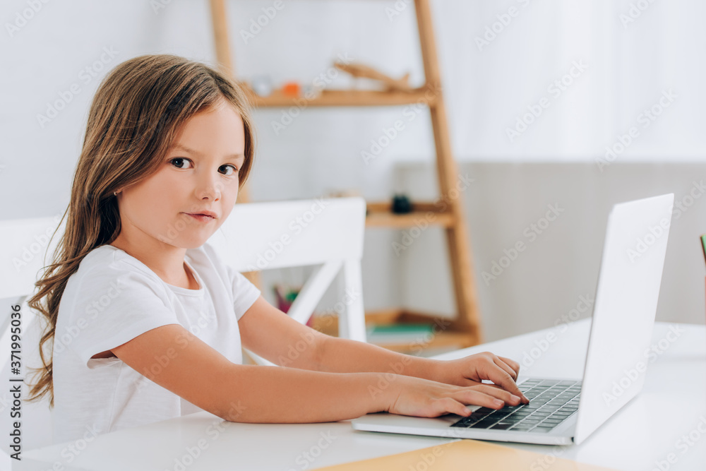 Obraz premium girl in white t-shirt looking at camera while sitting at table and using laptop