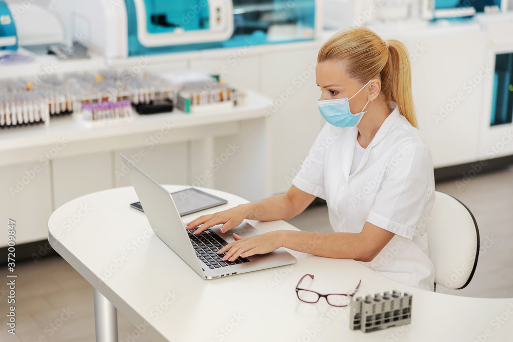 Beautiful blond female lab assistant sitting in lab with protective mask on and using laptop during covid outbreak. Beautiful blond female lab assistant sitting in lab with protective mask on and using laptop during covid outbreak.