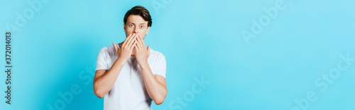 Panoramic orientation of shocked man covering mouth with hands on blue background