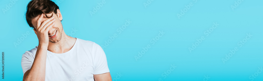 Panoramic crop of man covering eyes with hand isolated on blue Panoramic crop of man covering eyes with hand isolated on blue