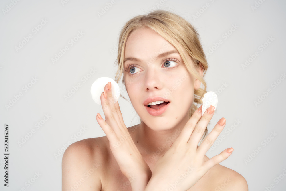 A blonde with bare shoulders holds cotton pads near her face skin care light background A blonde with bare shoulders holds cotton pads near her face skin care light background