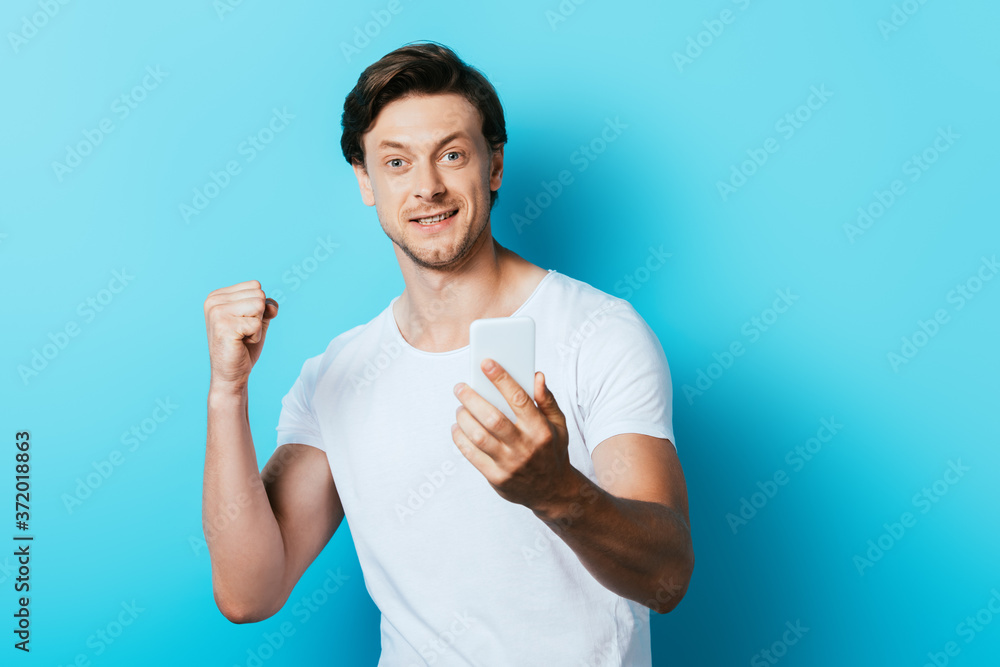Young man looking at camera and showing yeah gesture while holding smartphone on blue background Young man looking at camera and showing yeah gesture while holding smartphone on blue background
