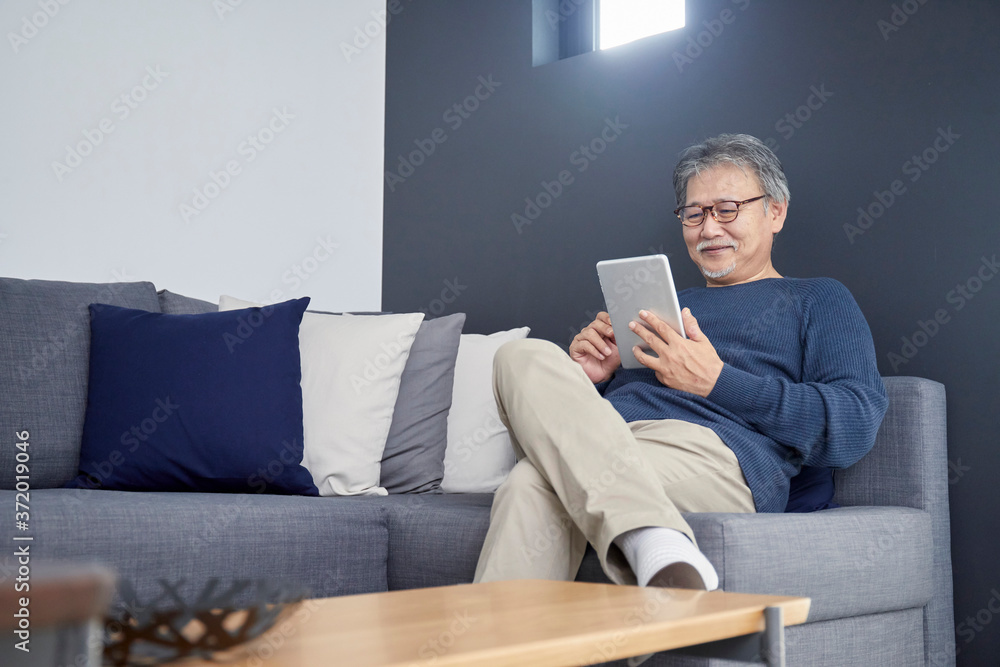 Fototapeta premium ソファーでくつろぎながらタブレットを操作するシニアの男性