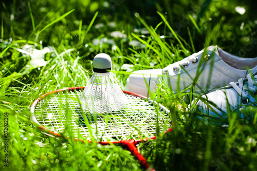 White shuttlecock, badminton racket andwhite sneakers lie on the green grass. Bright summer sunny day. Concept healthy lifestyle.