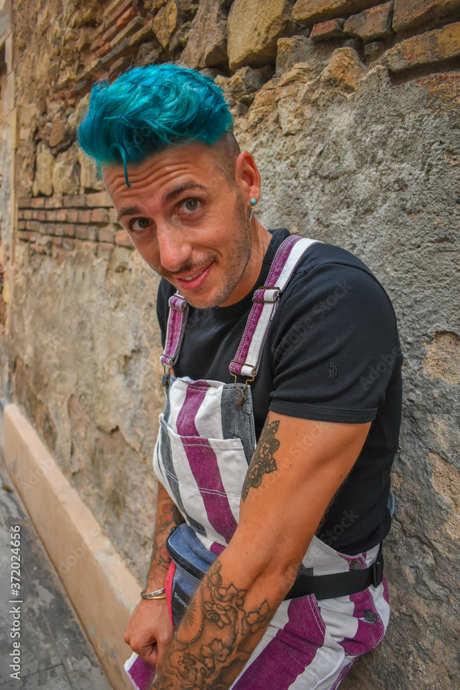 Young man wearing overalls, with blue hair, leaning against an old brick wall