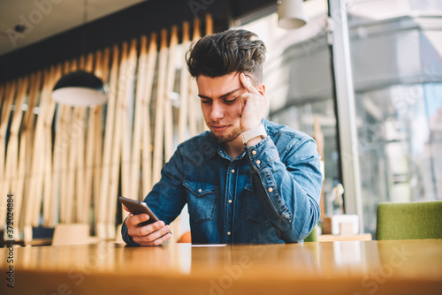 Stressed male feeling headache getting bill on smartphone email sitting at table in cafeteria, disappointed hipster guy reading bad news in income message on telephone having pain about trouble