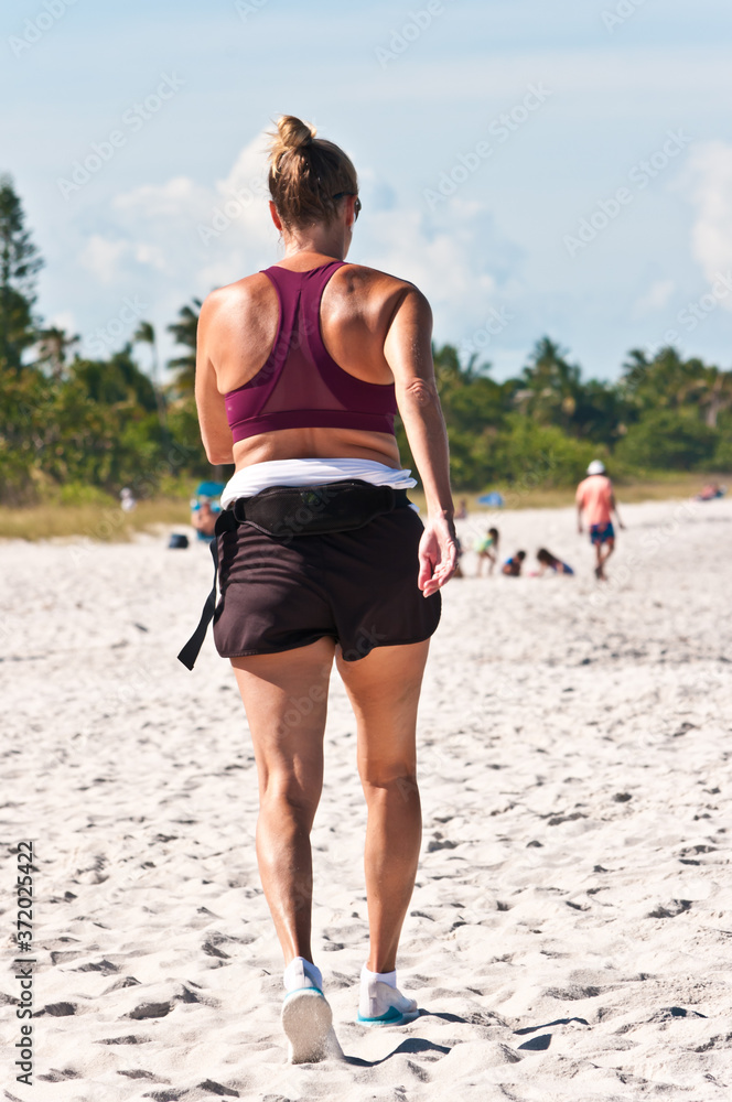 Back view, medium distance, of a middle aged woman briskly walking a tropical, sandy beach, on the gulf of Mexico on a sunny morning
