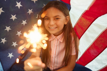 Portrait of cute girl holding sparkling lights smiling at camera against USA flag background, copy space