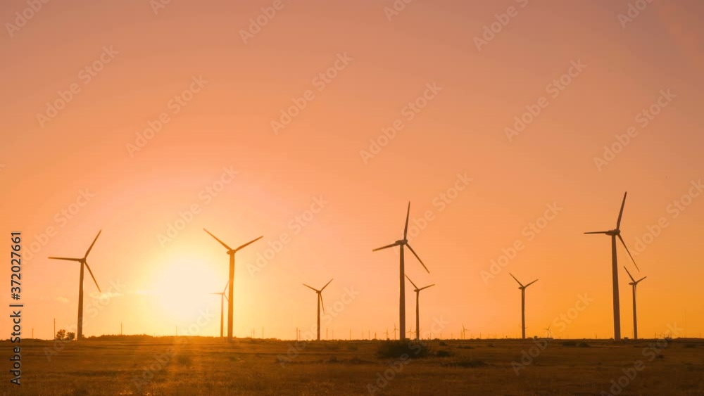 Spinning wind turbines in field against big sun at sunset with warm orange sky. Eco energy concept Spinning wind turbines in field against big sun at sunset with warm orange sky. Eco energy concept