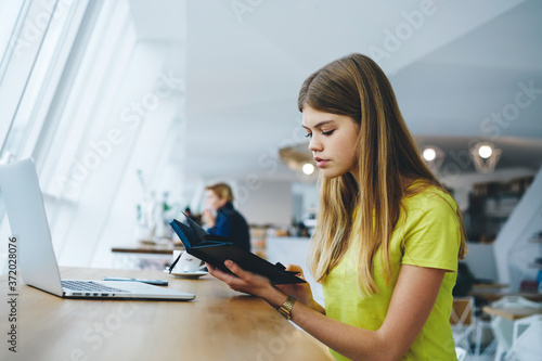 Intelligent hipster girl reading textbook sitting at digital computer device in coworking space.Smart female student with notebook in hand searching information in coffee shop indoors.Copy space area