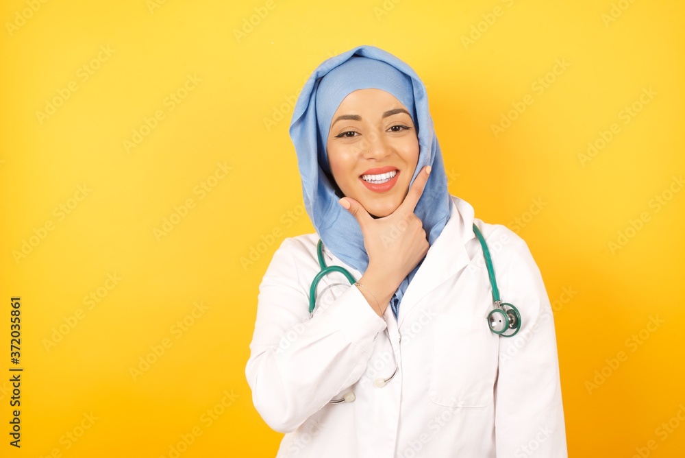Young arab doctor man wearing medical uniform standing over yellow background , looking confident at the camera smiling with crossed arms and hand raised on chin. Thinking positive. Young arab doctor man wearing medical uniform standing over yellow background , looking confident at the camera smiling with crossed arms and hand raised on chin. Thinking positive.