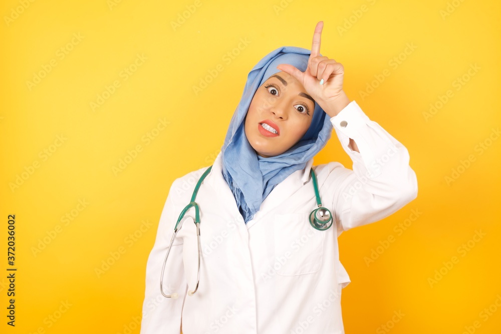 Φωτογραφία young beautiful Arab doctor woman wearing medical uniform standing over yellow background making fun of people with fingers on forehead doing loser gesture mocking and insulting