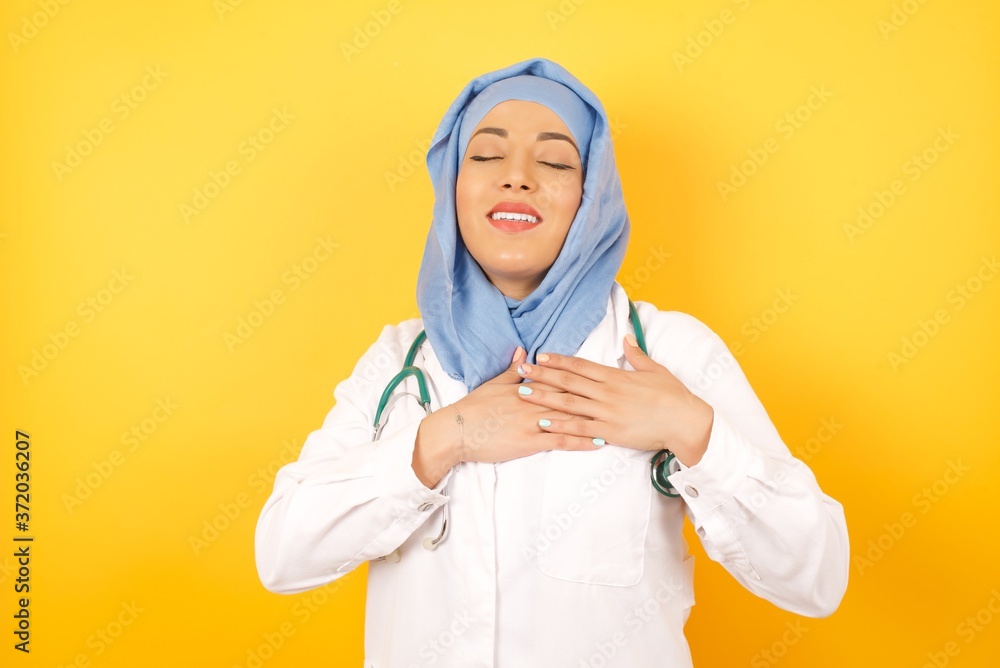 young beautiful Arab doctor woman wearing medical uniform standing over yellow background smiling with hands on chest with closed eyes and grateful gesture on face. Health concept.
