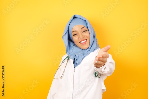 Young arab doctor woman wearing medical uniform standing over yellow background  smiling friendly offering handshake as greeting and welcoming. Successful business. © Roquillo