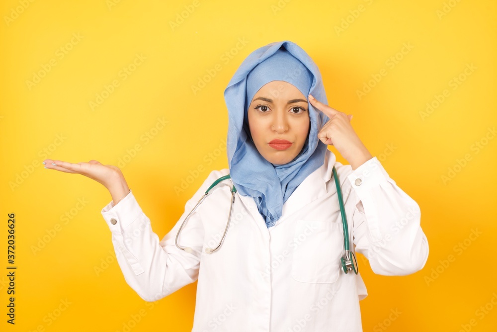 Young arab doctor woman wearing medical uniform standing over yellow background confused and annoyed with open palm showing copy space and pointing finger to forehead. Think about it. Young arab doctor woman wearing medical uniform standing over yellow background confused and annoyed with open palm showing copy space and pointing finger to forehead. Think about it.