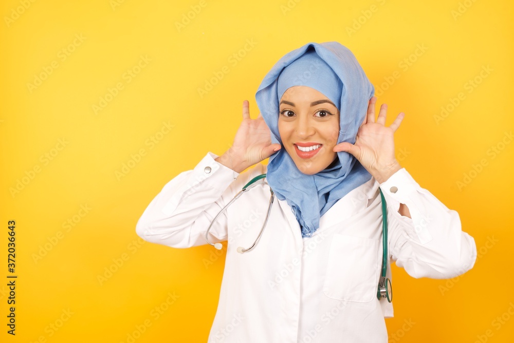 Φωτογραφία Young arab doctor woman wearing medical uniform standing over yellow background  Trying to hear both hands on ear gesture, curious for gossip