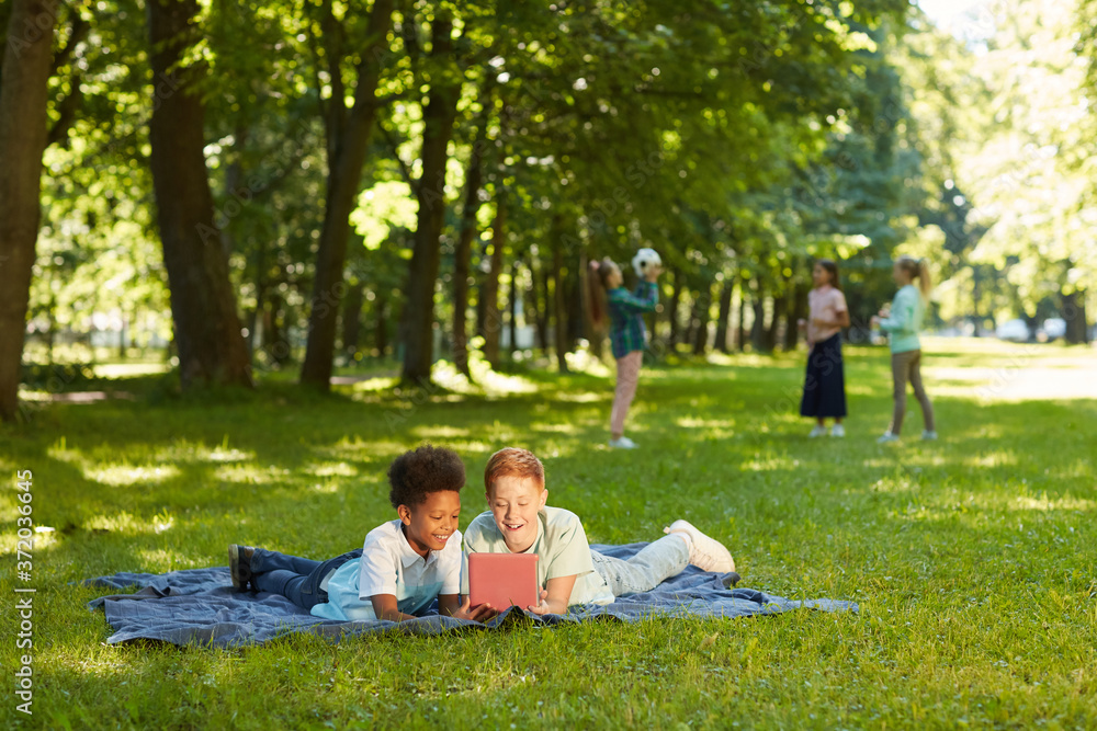 Wide angle portrait of two cheerful boys using digital tablet while lying on green grass in park outdoors lit by sunlight, copy space Wide angle portrait of two cheerful boys using digital tablet while lying on green grass in park outdoors lit by sunlight, copy space