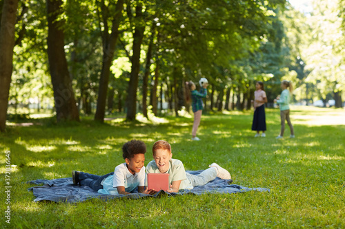 Wide angle portrait of two cheerful boys using digital tablet while lying on green grass in park outdoors lit by sunlight, copy space