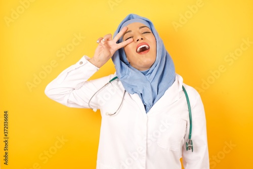 Young arab doctor woman wearing medical uniform standing over yellow background  Doing peace symbol with fingers over face  smiling cheerful showing victory